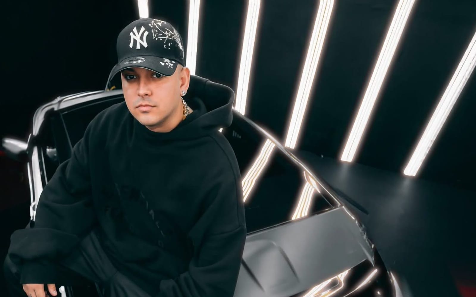 DJ Tao con gorra de Nueva York junto a un auto, entre luces blancas
